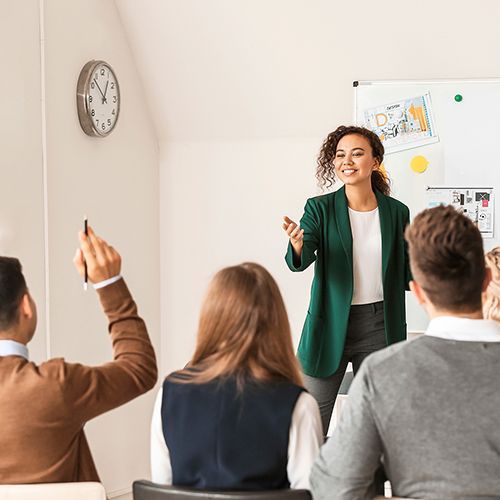 In einem Seminarraum sitzen Teilnehmende, eine Person meldet sich. Eine weibliche Lehrkraft steht an einem Whiteboard, das an der Wand hängt.