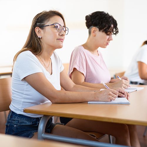Zei Teilnehmde sitzen in einem Seminarraum. Im Fokus ist eine junge Frau mit Brille, die einer Person außerhalb des Bildes zuhört. Sie hat einen Kugelschreiber in der Hand und ein Notizbuch vor sich liegen. Die anderen Frau hat ebenfalls Schreibgerät und Hefte vor sich liegen.