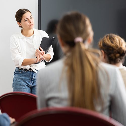 Eine junge Frau steht mit einer Mappe vor Publikum und spricht. Alle hören ihr aufmerksam zu. Im Hintergrund ist ein schwarzer Bildschirm.