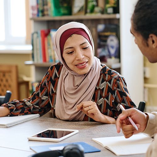 An einem Tisch sitzen drei People of Colour. Eine Frau mit Kopftuch beugt sich zu einem Mann und schaut auf dessen Unterlagen und er erklärt ihr etwas. Auf dem Tisch liegen Blöcke und ein Tablet