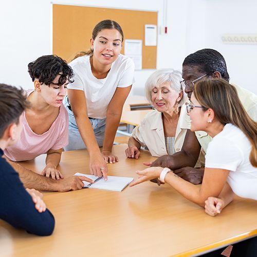 Eine Gruppe von unterschiedlichen Personen beugen sich über einen Zettel der in der Mitte auf einem Tisch liegt und diskutieren.