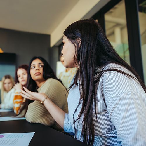 In einem Besprechungsraum sitzen mehrere Personen an einem langen dunklen Tisch. Sie sind von schräg unten aufgenommen. 
Im Fokus ist eine junge Frau mir langen schwarzen Haaren und einer gestreiften Bluse.  die zu den anderen hinhiseht, deswegen sieht man ihr Gesicht nicht richtig.