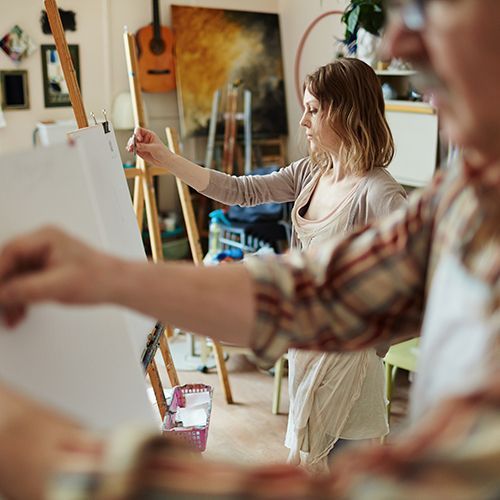 In einem Atelier stehen mehrere Staffeleien. Ein Mannzeichnet auf eine Staffelei im Vordergrund, er ist unscharf, Eine Frau an einer weiteren Staffelei steht konzentriert davor und hält einen Stift vor ihr.