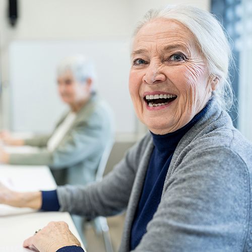 Eine ältere Frau sitzt in einem Seminarraum und lacht zur Seite in die Kamera. Im Hintergrund sind weitere ältere Personen unscharf zu erkennen.