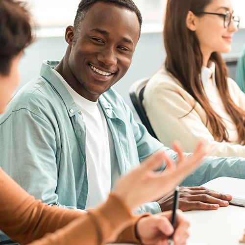 Vier junge Menschen sitzen nebeneinader an einem Tisch. Im Vordergrund unterhalten sich zwei Männer, einer lacht den anderen an, den man nur von hinten sieht. Die beiden Frauen danaben schauen in ihr Tablet.