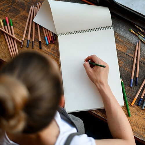 Auf einer breiten, dunklen Holzfensterbank liegt ein Skizzenblock. Eine Frau hält einen grünen Stift in der Hand. Die Perspektive ist von oben auf den Block.