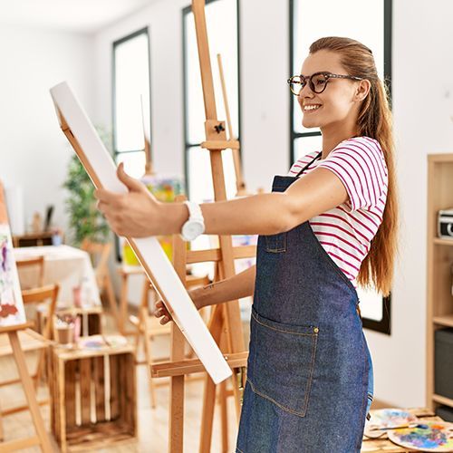 Eine lachende junge Frau begutachtet in einem Atelier ihr Bild auf einer Leinwand. Auf einer Staffelei ist ein weiteres Bild zu sehen. Ebenso ein Regal mit dunklen Kisten