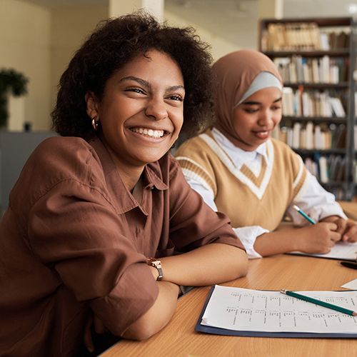 Drei Menschen of Colour sitzen an einem Tisch voller Papier, einem Laptop, einem Klemmbrett und Stiften. Im Hintergrund ist ein Bücherregal zu sehen. Im Fokus ist eine junge Frau die lachend an der Kamera rechts vorbeischaut.