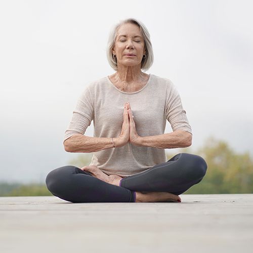 Eine ältere Frau sitzt auf einem grauen Boden draußen im Lotussitz. Sie hat die Augen gescjlossen und die Hände vor der BNrust aneinandergelegt. Im Hintergrund sind Baumkronen auf der Höhe des Bodens unscharf zu sehen.