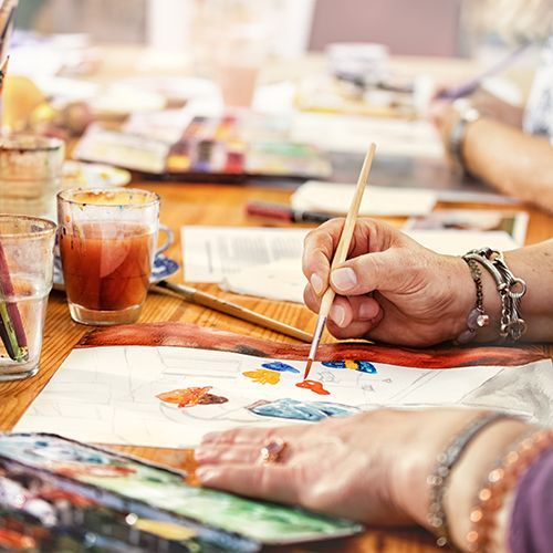 Zwei Frauen mittleren Alters malen mit Aquarellfarben Bilder. Man sieht, wie sie einen Pinsel in der Hand halten und das Blatt vor ihnen kolorieren. Auf dem Tisch stehen viele Materialien, wie z.B. Farbkästen und Behälter mit einer großen Auswahl an Pinsel.