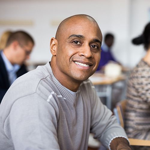 Ein Man of Colour schaut lächelnd in die Kamera. Im Hintergrund sind weitere Personen zu sehen die an Tischen sitzen und Stifte und Papier vor sich liegen haben.