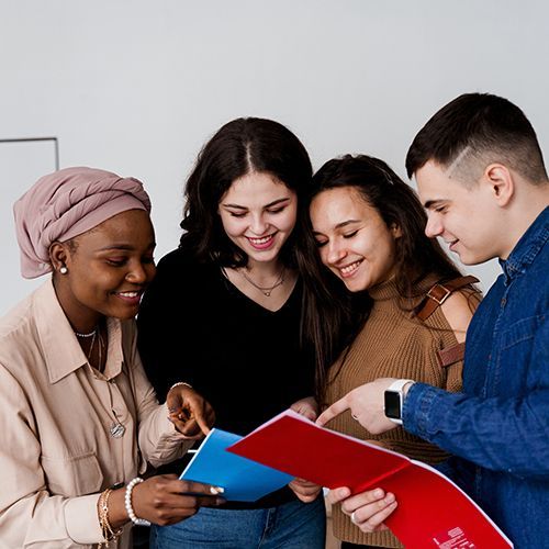Eine Gruppe junger People of Colour stehen in einem Raum und schauen gemeinsam in ein blaues und ein rotes Heft. Sie zeigen sich dabei etwas. Alle lächeln.