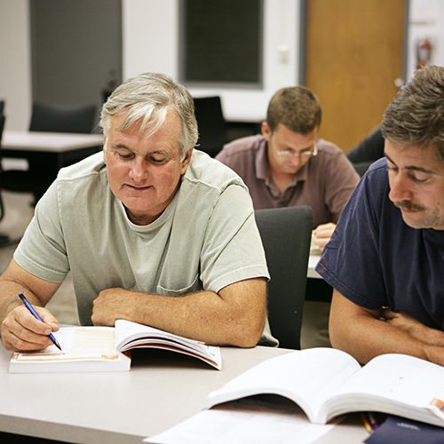 Männliche Teilnehmende sitzen in einem Seminarraum zu zweit an Tischen . im Fokus sind zwei Männer zu sehen, die in ihr Buch schauen, das vor ihnen liegt. Ein Mann zeigt mit einem Stift ins Buch.