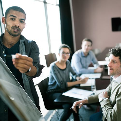 Ein Mann präsentiert an einem Flip-Chart einer Gruppe von Menschen, die um einen Tisch sitzen etwas. Im Hintergrund ist eine matte, lilafarbene Wand mit einem Bildschirm zu sehen.