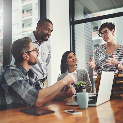 Eine Gruppe von Personen sitzt und steht an einem Holztisch und redet miteinander. Auf dem Tisch steht ein Laptop im Hintergrund sind hohe Fenster.