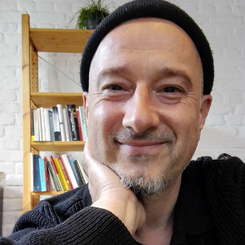 Portrait eines Mannes mit Mütze und Bart, der lächelnd vor einem Bücherregal sitzt.